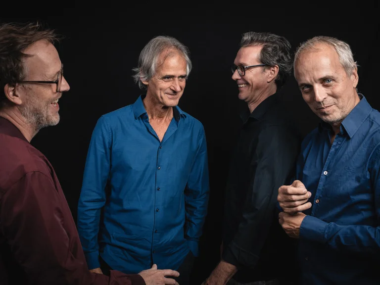 Vier Männer (Markus Stockhausen's QUADRIVIUM) stehen entspannt zusammen. Sie tragen dunkle Hemden und lächeln in einem Studio mit dunklem Hintergrund.