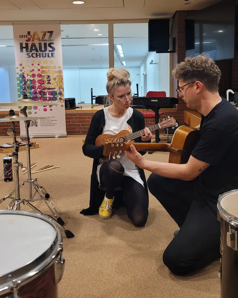 Zwei Personen knien in einem Musikraum, wo eine Frau Ukulele spielt und ein Mann mit Gitarre dabei hilft.
