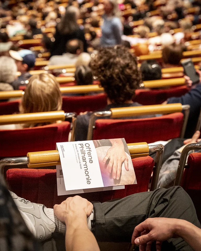 Ein Publikum sitzt in einem Konzertsaal (die Kölner Philharmonie). Im Vordergrund hält jemand ein Programmheft der Offene Philharmonie auf dem Schoß.
