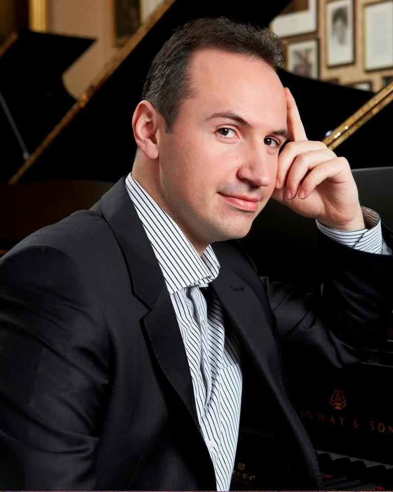 Simon Trpčeski: Ein Mann in einem Anzug sitzt lächelnd neben einem Flügel. Der Raum im Hintergrund enthält gerahmte Bilder an der Wand.