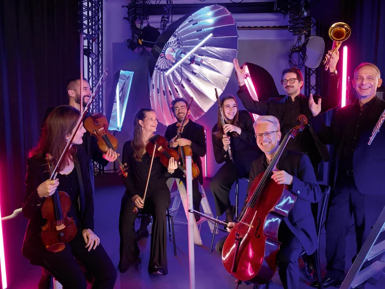 Ein fröhliches Orchester mit acht Musikern in einem farbenfrohen Raum spielt verschiedene Saiten- und Blechblasinstrumente. Beleuchtung und Dekor sind modern.