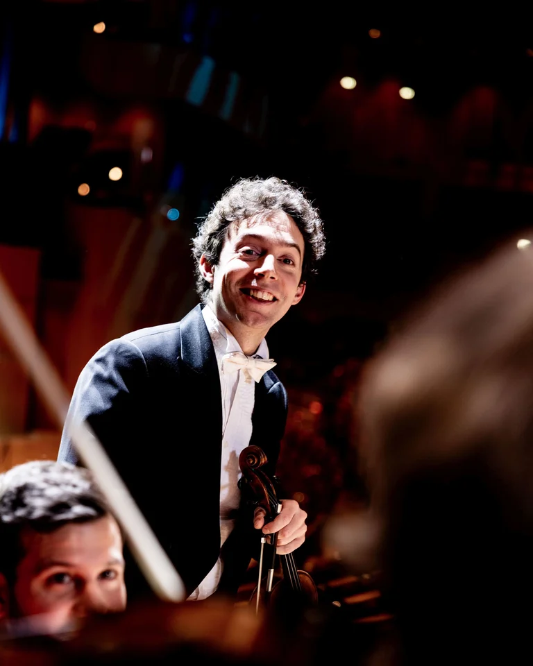 Ein lächelnder Dirigent in Frack steht vor einem Orchester. Die warme Beleuchtung schafft eine festliche Atmosphäre.