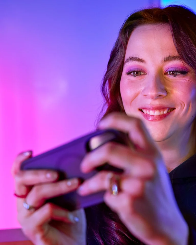 Eine Person hält ein Smartphone, lächelt, vor einem farbenfrohen Hintergrund mit rosa und blauem Licht.