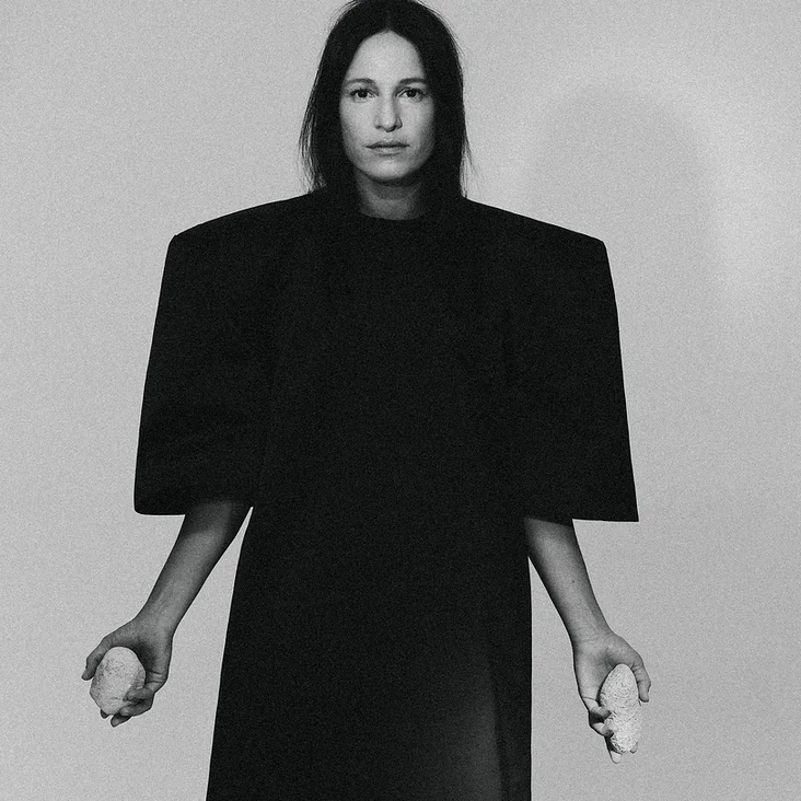 Schwarz-weißes Porträt einer Person (Carminho) in einem langen, dunklen Gewand mit breiten Schultern, die zwei weiße Gegenstände hält.