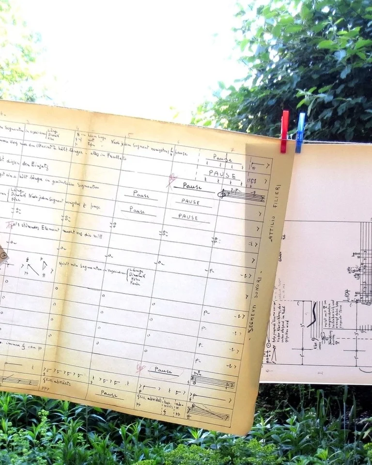 Pläne und Zeichnungen hängen an einer Wäscheleine im Freien. Sie sind mit bunten Klammern befestigt und von Bäumen umgeben.