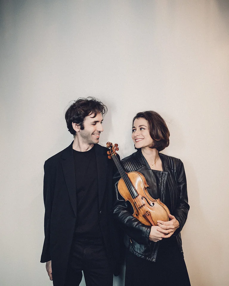 Ein Mann und eine Frau (Alexandre Kantorow und Liya Petrova) in schwarzer Kleidung lächeln einander an. Die Frau hält eine Geige in der Hand.