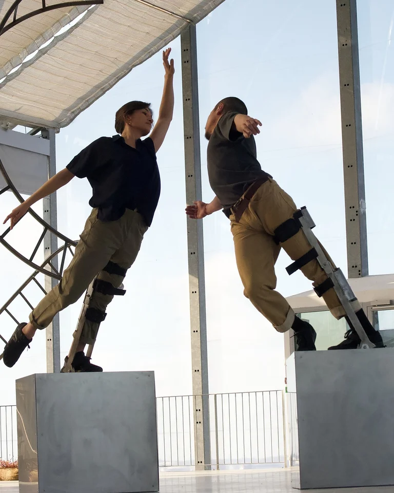 Zwei Tänzer auf Podesten führen eine akrobatische Performance vor. Sie tragen spezielle Beinverlängerungen und sind in einem modernen Raum.