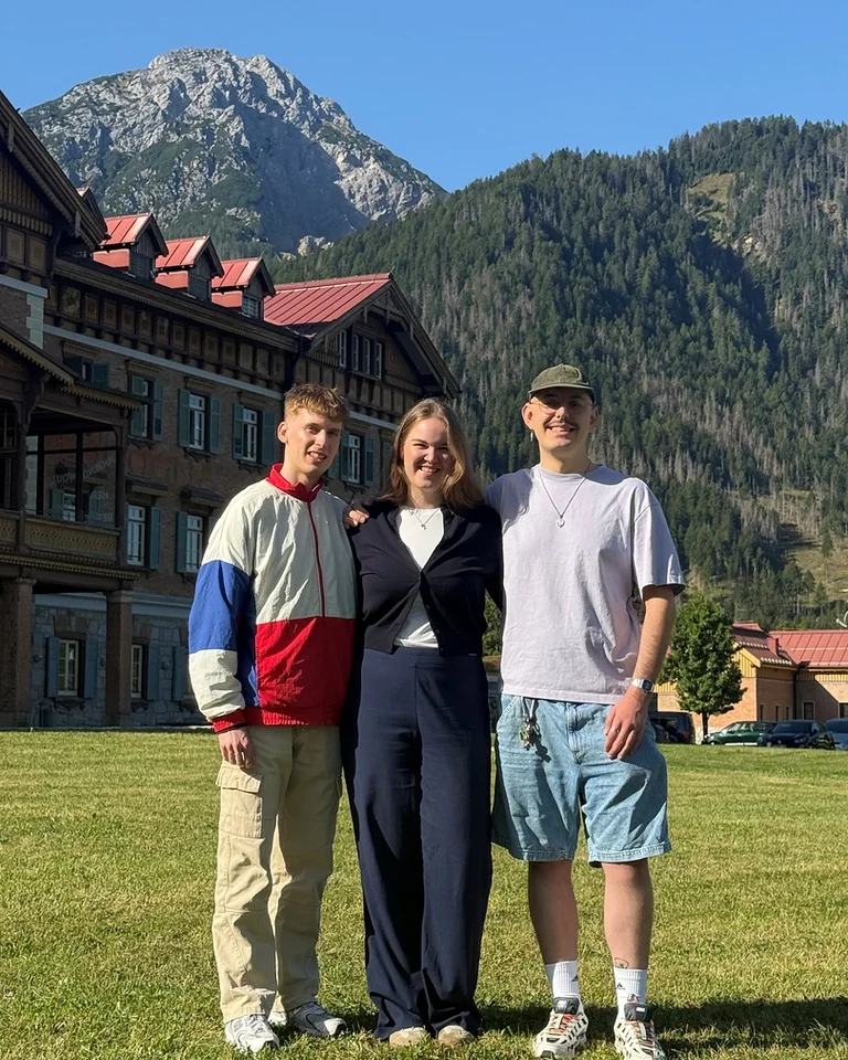 Drei Personen (Mahler Ambassadors) stehen lächelnd in einer grünen Wiese vor einem großen Gebäude mit Bergen im Hintergrund an einem klaren Tag.