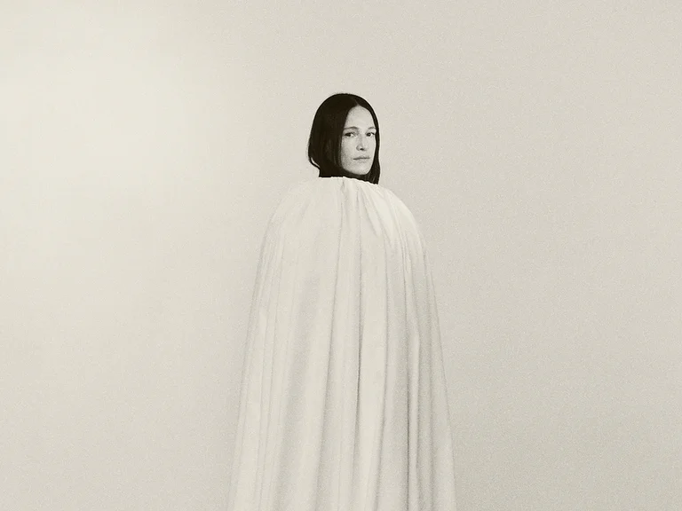 Eine Frau (Carminho) steht still, in ein langes, weißes Tuch gehüllt, mit ernstem Ausdruck. Minimalistischer Hintergrund. Schwarz-weiß Fotografie.