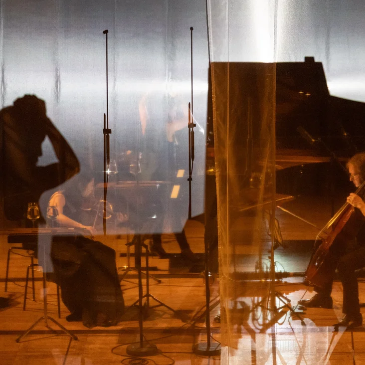 Ein Cellist spielt auf einer Bühne mit gespiegelten Silhouetten anderer Musiker. Die Atmosphäre ist warm und konzentriert.