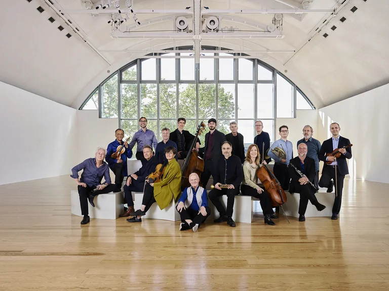 Eine Gruppe von 16 Musikern (Ensemble Modern) posiert in einem modernen, lichtdurchfluteten Raum mit großen Fenstern und einem Holzboden.
