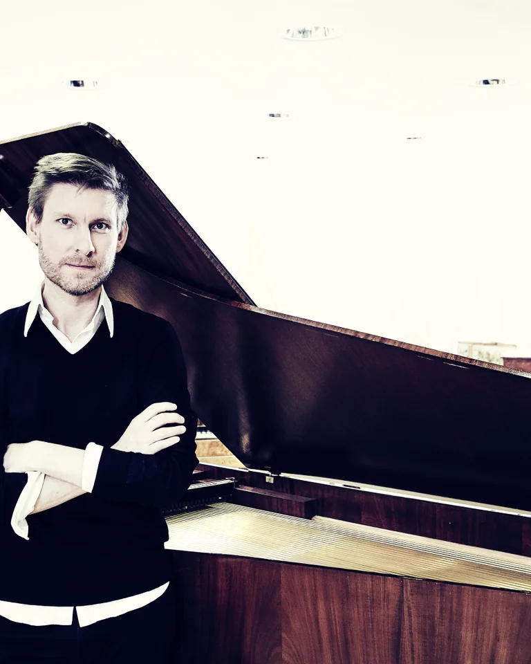 Eine Person steht mit verschränkten Armen vor einem Flügel in einem minimalistischen Raum. Die Atmosphäre ist modern und klar.