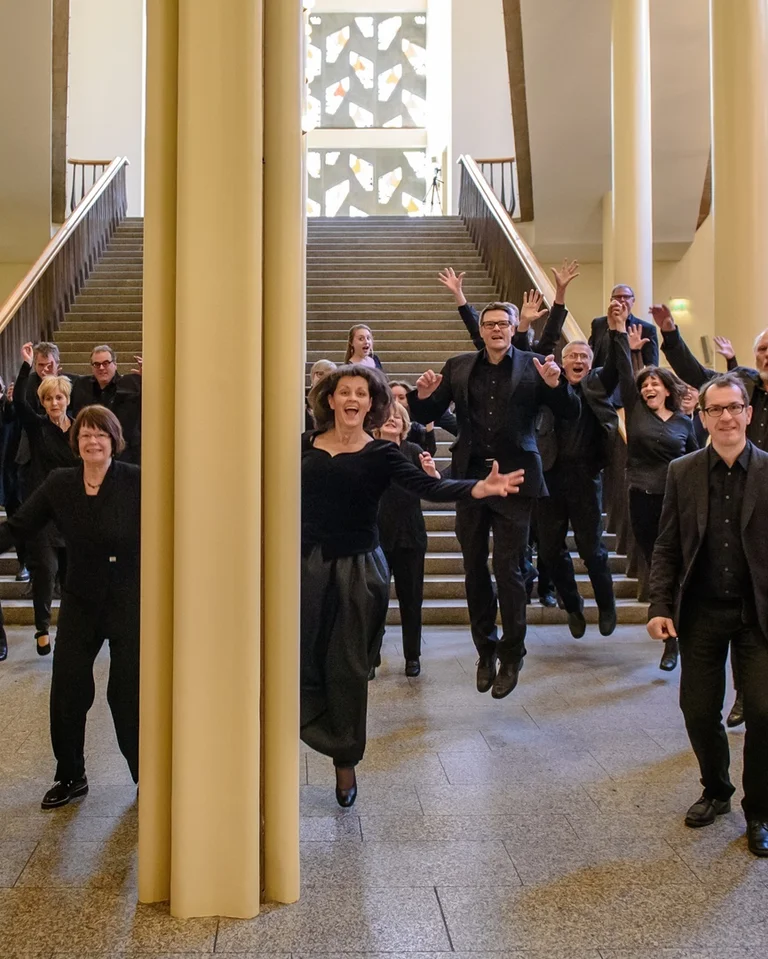 Eine Gruppe von Menschen (Gürzenich-Chor Köln), alle in Schwarz gekleidet, springt freudig in einer großen Halle mit Treppen im Hintergrund.