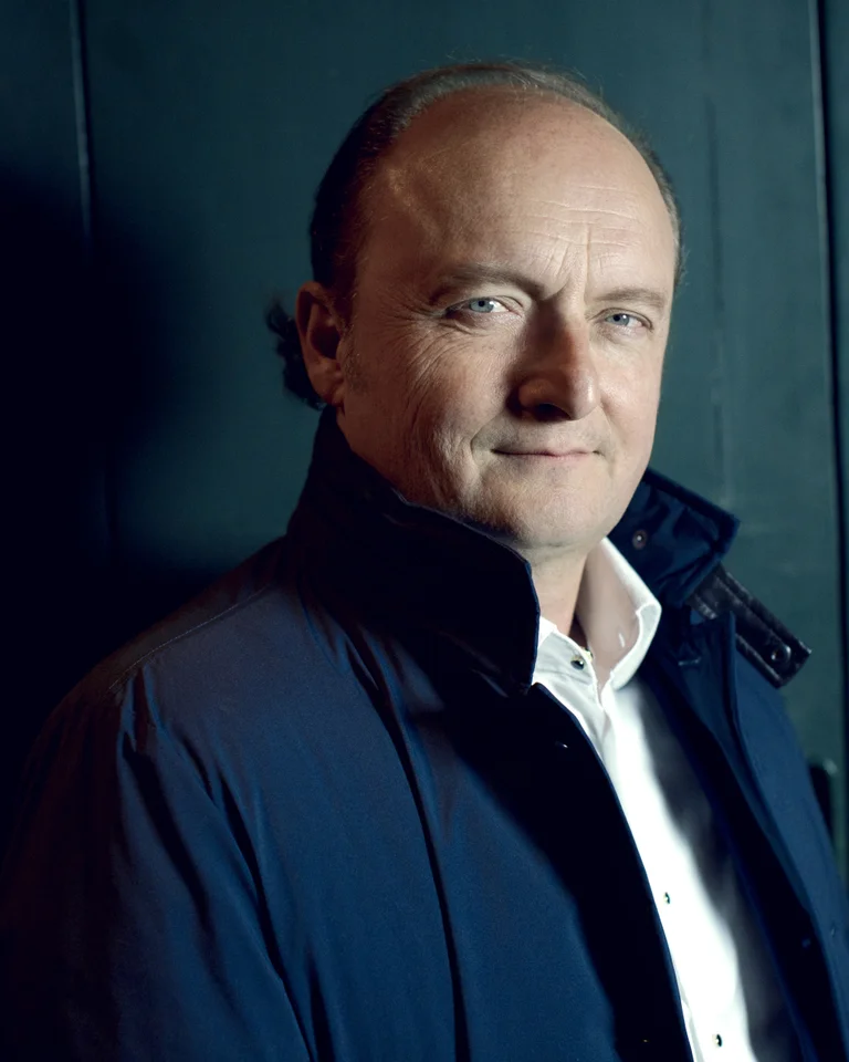 Ein Mann in einem dunkelblauen Mantel steht vor einem dunklen Hintergrund, schaut direkt in die Kamera.