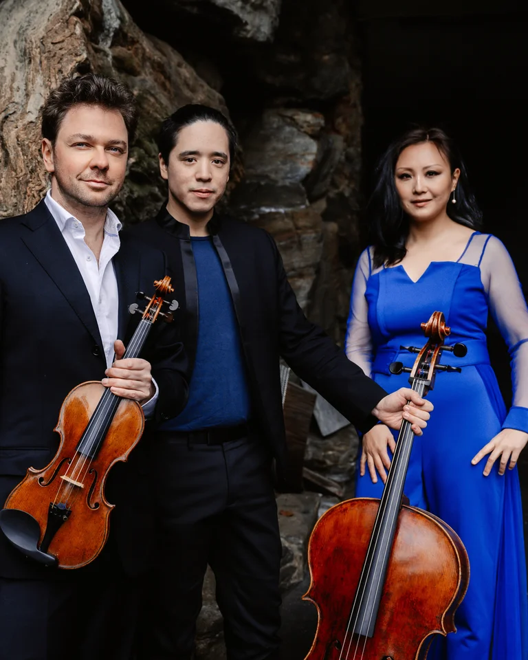 Drei Musiker posieren vor einem Felsen. Zwei halten Streichinstrumente, die dritte Person trägt ein elegantes, blaues Kleid.