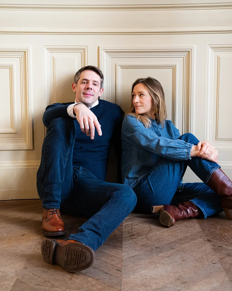 Ein Mann und eine Frau (Mathieu Pordoy und Sabine Devieilhe) sitzen entspannt auf einem Holzboden vor einer cremefarbenen Wand. Beide tragen Jeans.