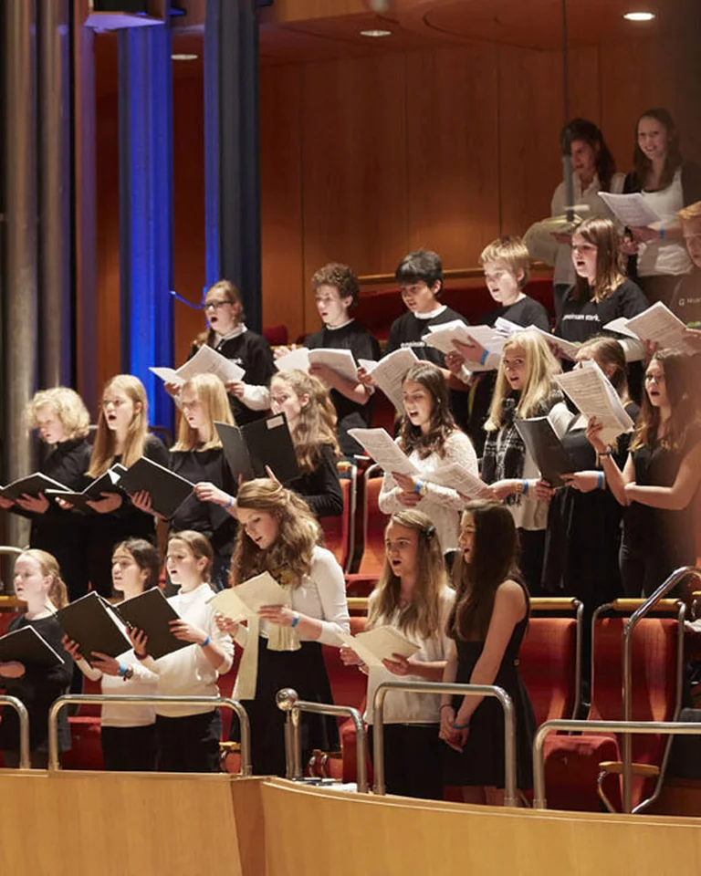 A choir performs in a concert hall with large organ pipes in the background. Singers hold sheet music in their hands.