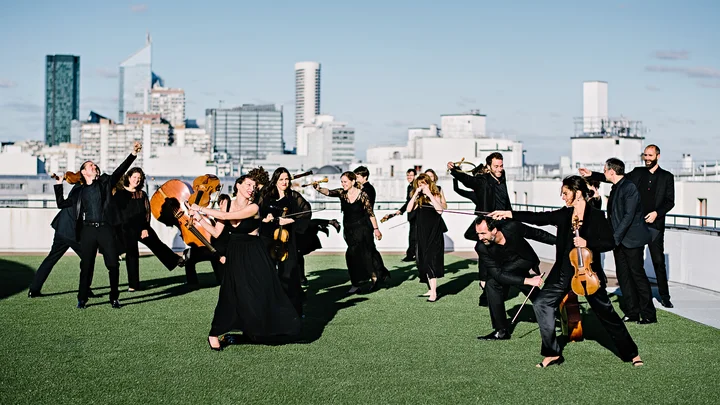 Eine Gruppe Musiker (Le concert de la Loge) in schwarzen Outfits spielt klassische Instrumente auf einem Dach vor einer städtischen Skyline bei klarem Himmel.