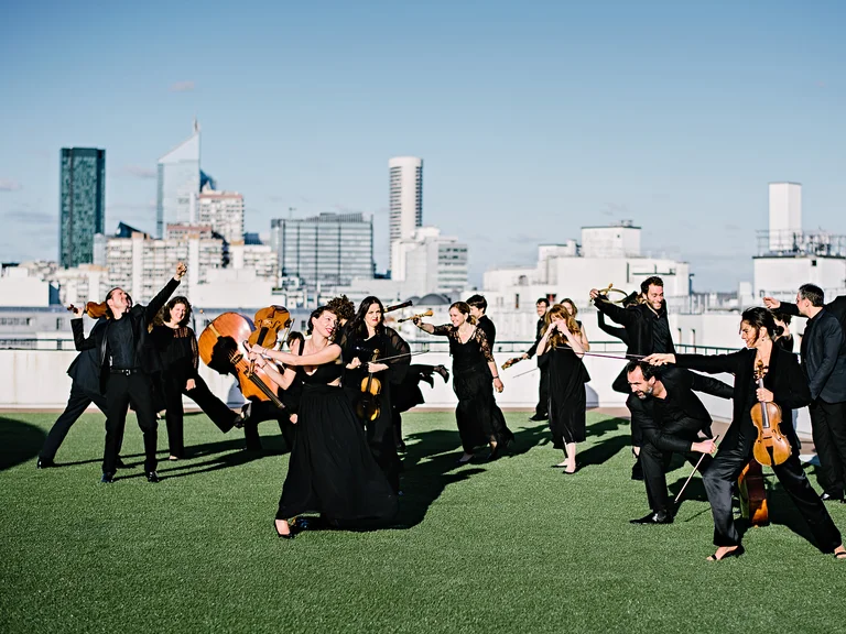 Eine Gruppe Musiker (Le concert de la Loge) in schwarzen Outfits spielt klassische Instrumente auf einem Dach vor einer städtischen Skyline bei klarem Himmel.