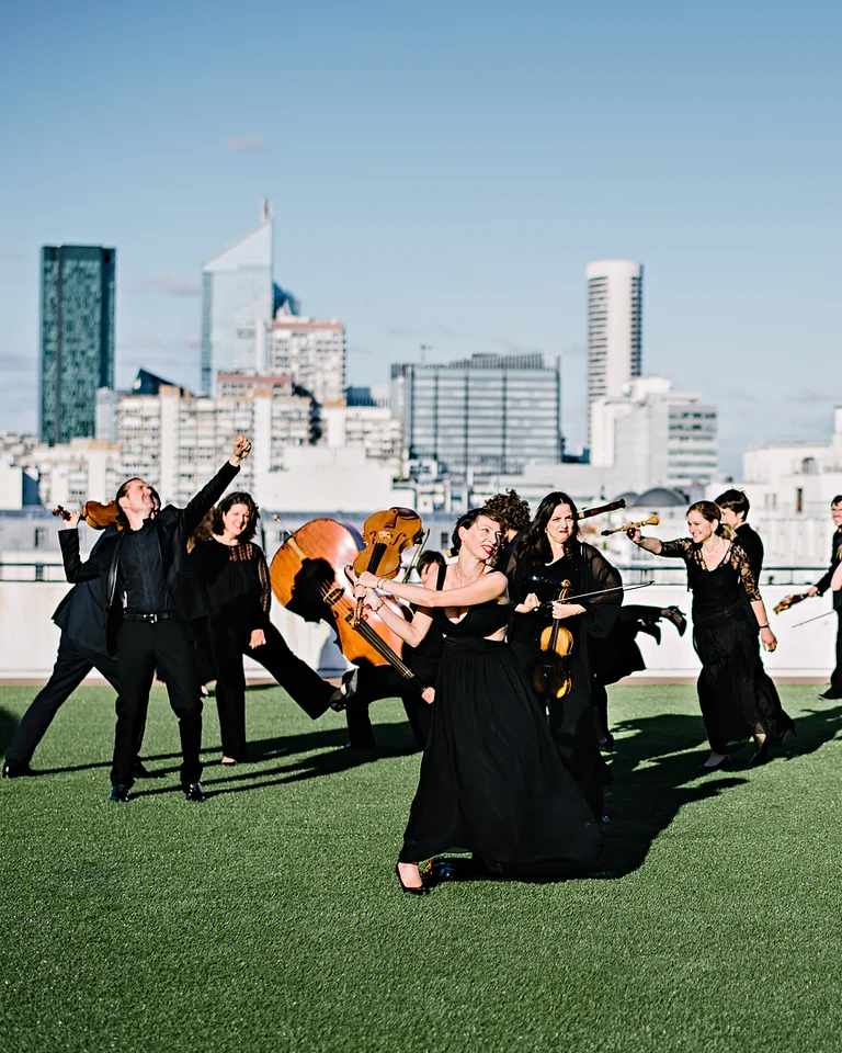 Eine Gruppe Musiker (Le concert de la Loge) in schwarzen Outfits spielt klassische Instrumente auf einem Dach vor einer städtischen Skyline bei klarem Himmel.
