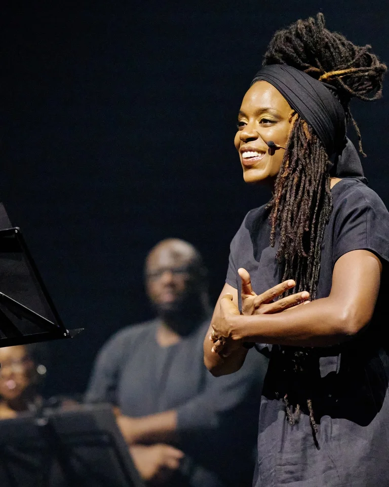 Akua Naru performs on a stage with a music stand in front of her, while two people watch in the background.
