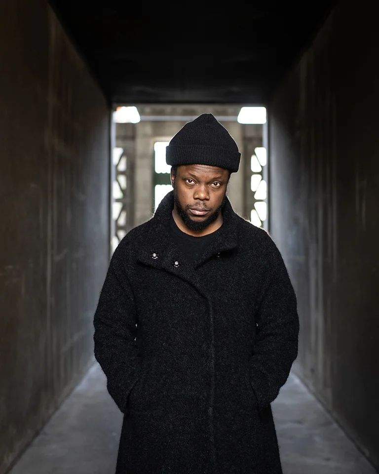 Ein Mann (Ambrose Akinmusire) steht in einem dunklen Korridor, trägt einen schwarzen Mantel und eine Mütze. Licht strahlt im Hintergrund.