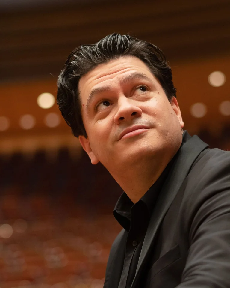 Eine Person im schwarzen Anzug sitzt in einem Konzertsaal und schaut nach oben, vor einem unscharfen Hintergrund.