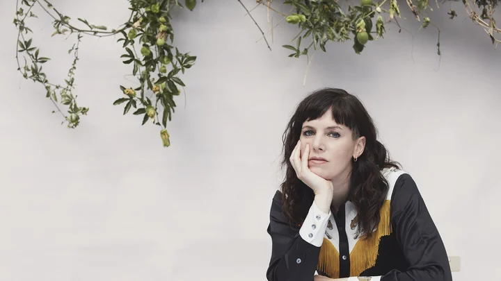 Eine Frau (Anna Meredith) in schwarz-weißem Hemd mit gelben Details sitzt an einem Tisch und stützt ihren Kopf in die Hand. Zweige im Hintergrund.