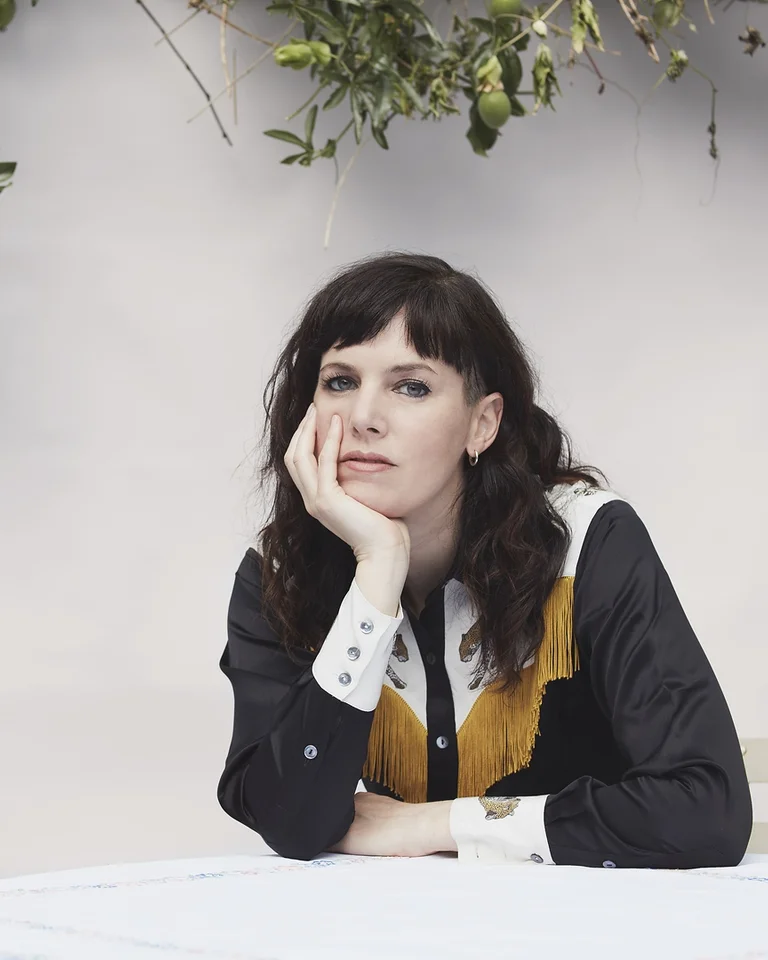 Eine Frau (Anna Meredith) in schwarz-weißem Hemd mit gelben Details sitzt an einem Tisch und stützt ihren Kopf in die Hand. Zweige im Hintergrund.