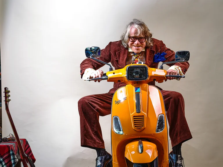 Ein Mann in einem braunen Anzug sitzt auf einem orangefarbenen Roller in einem Studio mit einer Gitarre im Hintergrund.