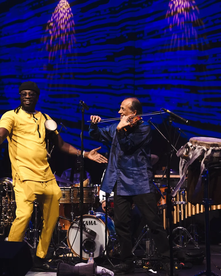 The musicians of the Deggo Band on stage, dressed in yellow, blue, and burgundy, flanked by drums and a blue backdrop. An energetic performance.