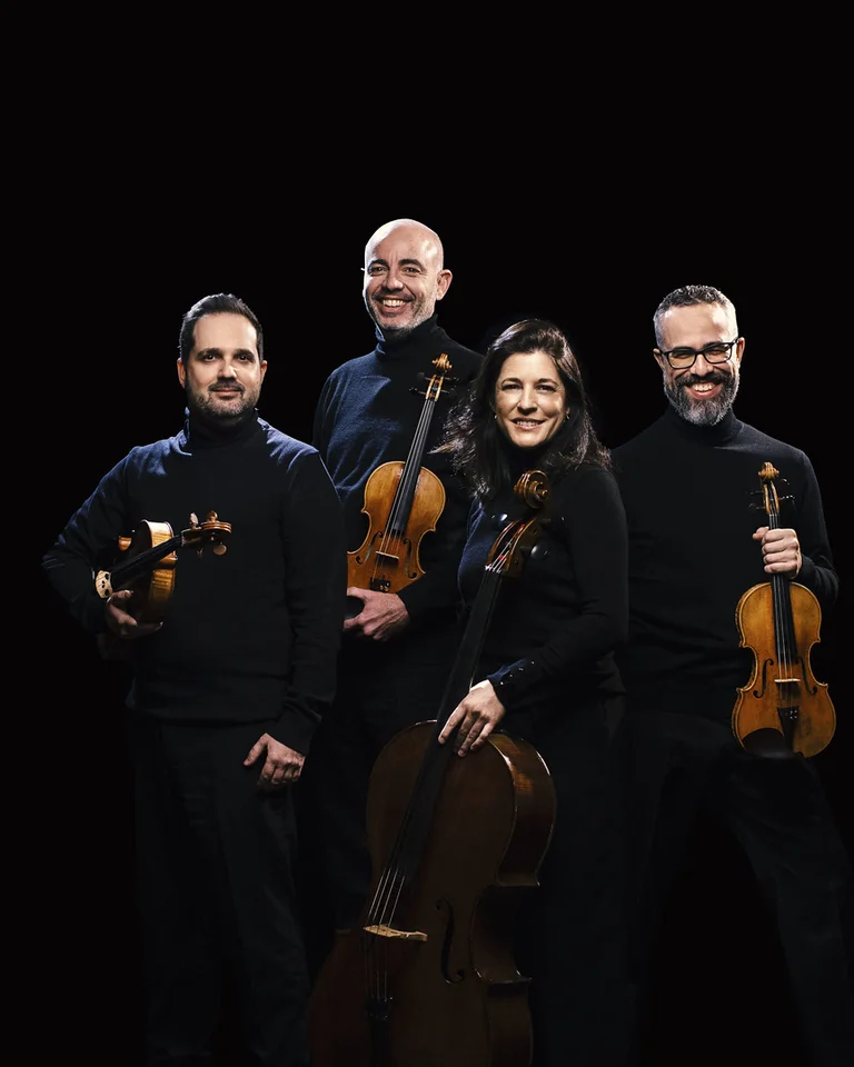Vier Musiker (Cuarteto Quiroga)posieren mit ihren Streichinstrumenten vor schwarzem Hintergrund. Alle tragen schwarze Kleidung und lächeln in die Kamera.