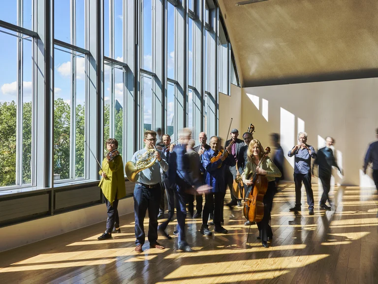 Eine Gruppe von Menschen (Ensemble Modern) versammelt sich in einem lichtdurchfluteten Raum mit großen Fenstern und Parkettboden. Sie betrachten aufmerksam etwas.