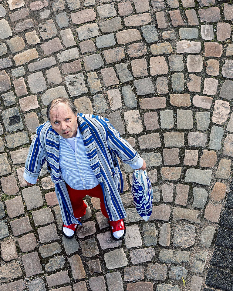 Ein älterer Mann steht auf einem Kopfsteinpflaster, trägt rotes und blaues Streifenoutfit. Perspektive von oben aufgenommen.