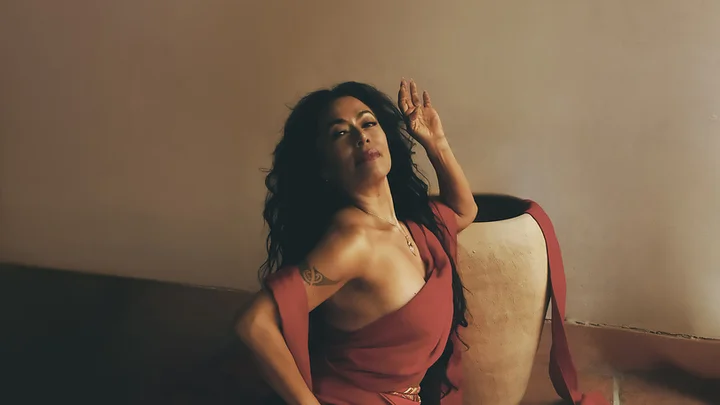 Eine Frau (Lila Downs) in einem roten Kleid posiert dramatisch auf dem Boden, umgeben von roten Stoffstreifen und einer großen Vase.