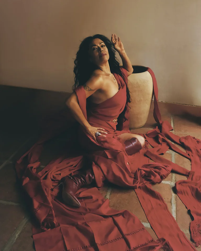 Eine Frau (Lila Downs) in einem roten Kleid posiert dramatisch auf dem Boden, umgeben von roten Stoffstreifen und einer großen Vase.