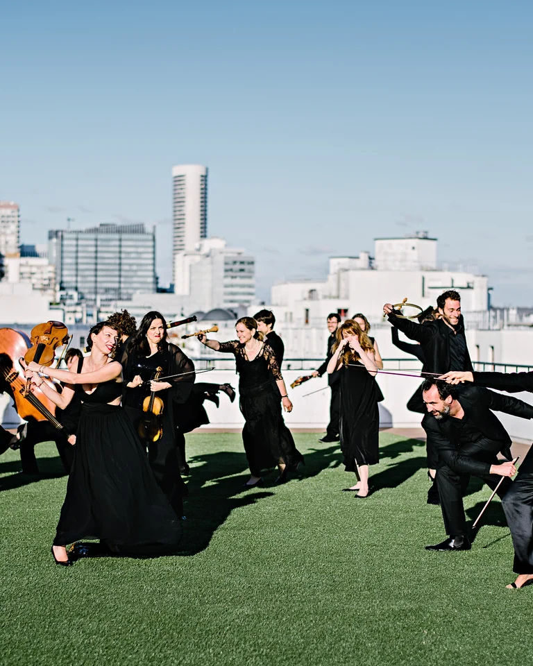 The members of the ensemble Le Concert de la Loge pose in elegant black attire, accompanied by several instruments, on a rooftop overlooking an urban skyline.