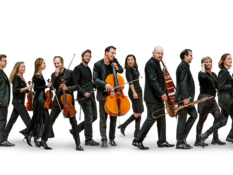 Gruppe von Musikerinnen und Musikern in formeller Kleidung mit ihren Instrumenten, darunter Violinen und Celli, schreitet im Gleichschritt.