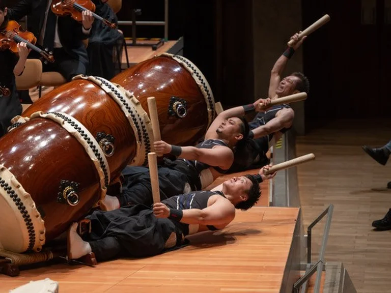 Vier Trommler (aus Waseda Symphony Orchestra Tokyo) spielen Taiko-Trommeln im Liegen auf einer Bühne. Sie tragen traditionelle Kleidung und halten große Trommelstöcke.