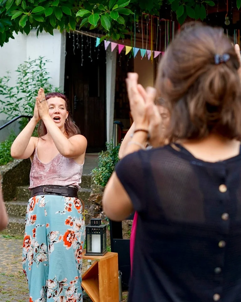 Gruppe von Menschen im Freien, die zusammen klatschen. Eine Frau in der Mitte (Clara Flaksman) lächelt, während andere sie umgeben. Farbenfrohe Dekorationen hängen im Hintergrund.