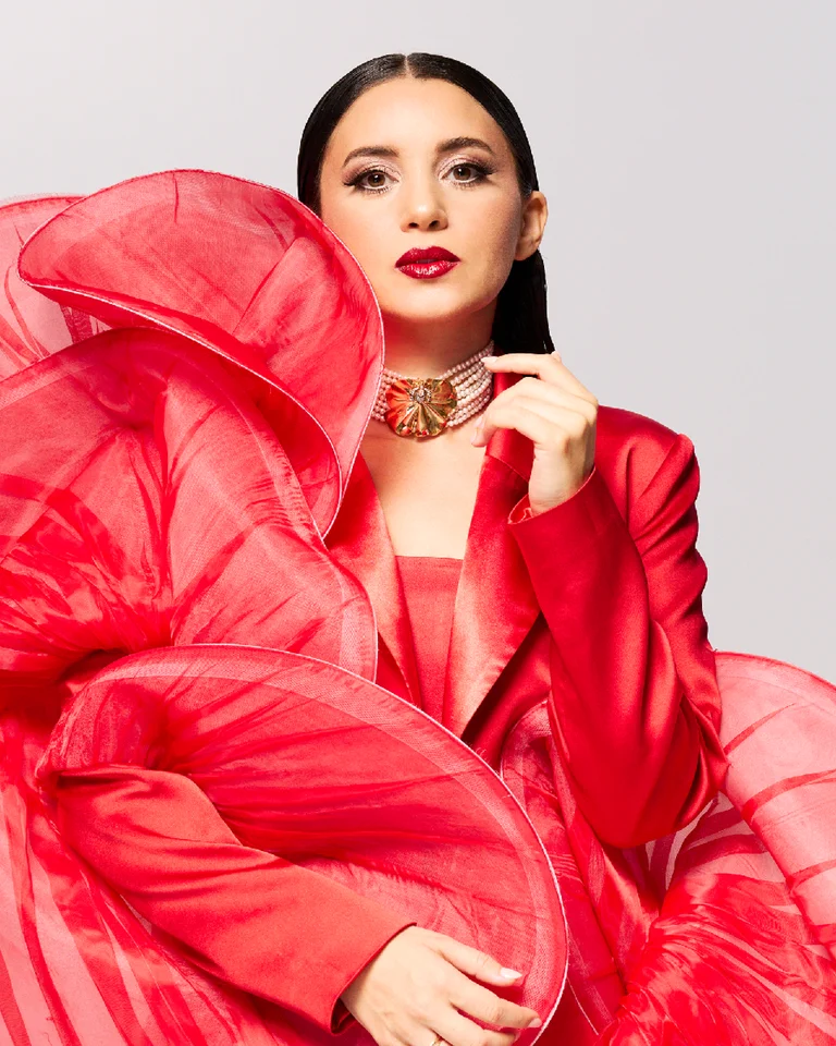 Eine Person (Karsu) in einem auffälligen roten Kleid mit vielen Rüschen posiert selbstbewusst vor einem schlichten, grauen Hintergrund.