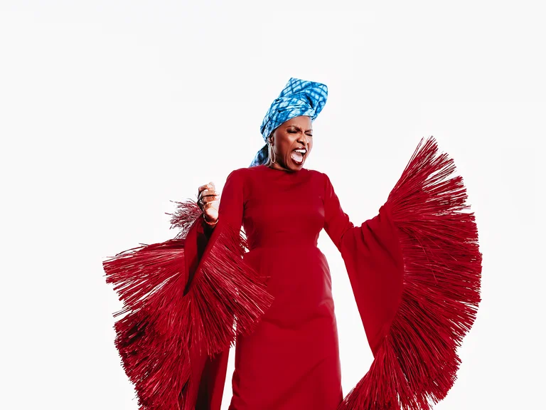 Frau (Angélique Kidjo) in fließendem rotem Gewand mit langen Fransenärmeln und blauem Kopftuch posiert energetisch vor hellem Hintergrund.