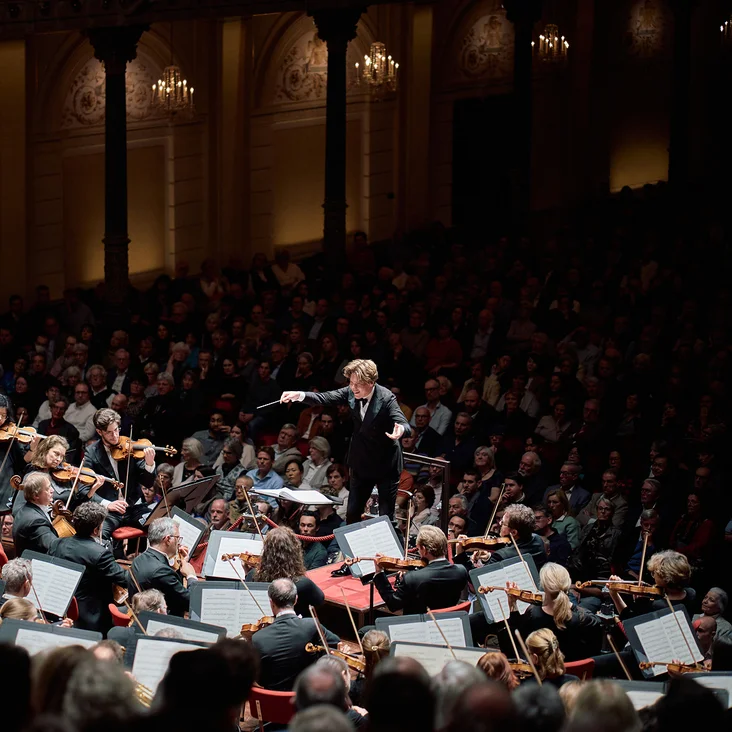 Ein großes Orchester (Royal Concertgebouw Orchestra) spielt in einem eleganten Konzertsaal. Der Dirigent (Klaus Mäkelä) steht im Vordergrund, das Publikum lauscht aufmerksam.