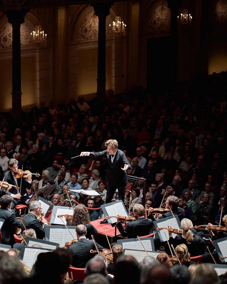 Ein großes Orchester (Royal Concertgebouw Orchestra) spielt in einem eleganten Konzertsaal. Der Dirigent (Klaus Mäkelä) steht im Vordergrund, das Publikum lauscht aufmerksam.