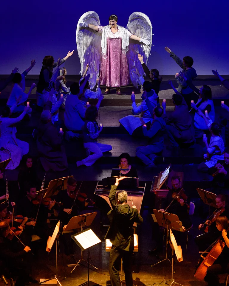 Eine Bühne mit einem Orchester im Vordergrund und einer Darstellerin im Engel-Kostüm im Hintergrund, dramatisch beleuchtet.