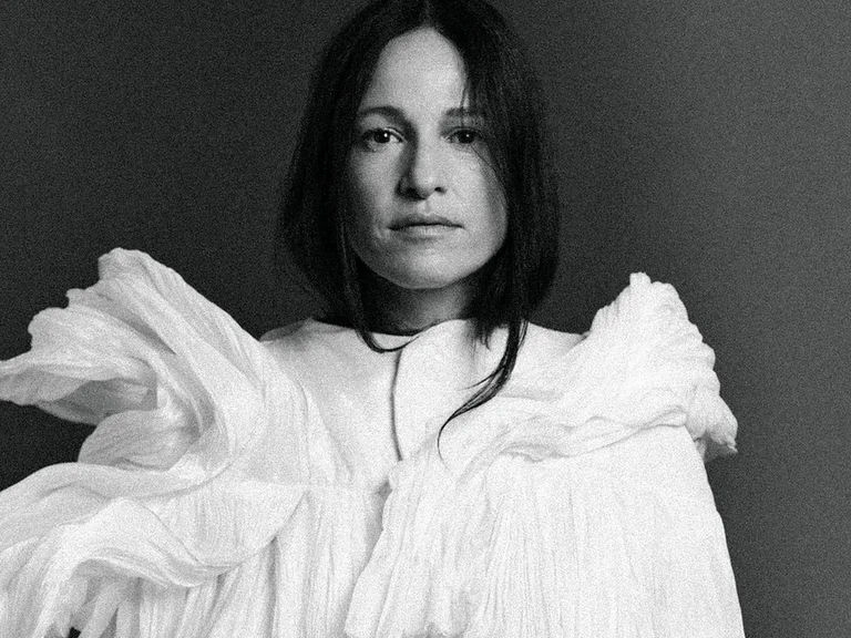 Eine Frau (Carminho) in einem weißen, kunstvoll drapierten Kleid, blickt ruhig in die Kamera. Der Hintergrund ist schlicht und dunkel.