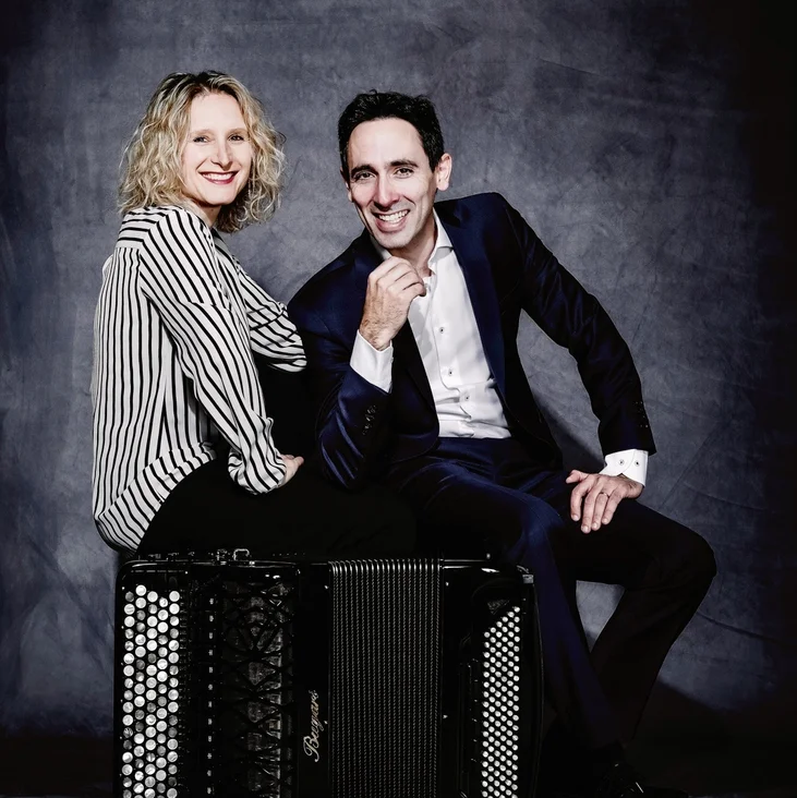 A man in a suit and a woman in a striped shirt (Marie-Andrée Joerger and Vincent Dubois) smiling, sitting or leaning on an accordion.