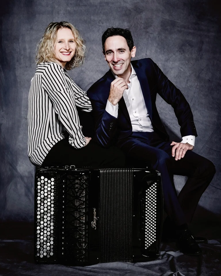 A man in a suit and a woman in a striped shirt (Marie-Andrée Joerger and Vincent Dubois) smiling, sitting or leaning on an accordion.
