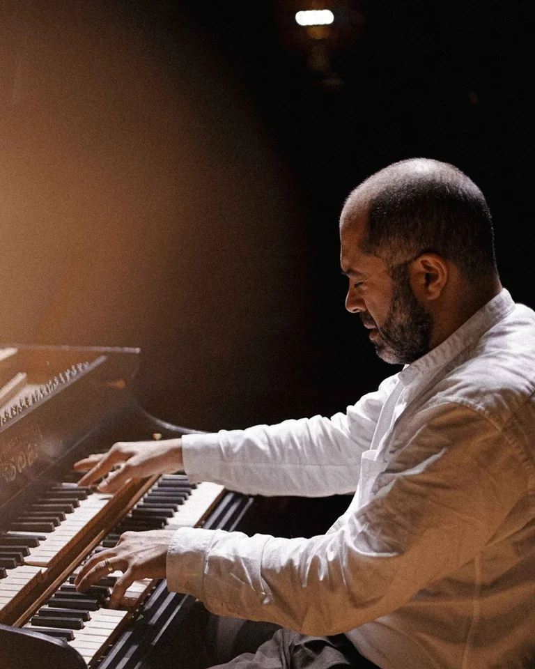 Ein Mann (Mahan Esfahani) in einem weißen Hemd spielt Cembalo auf einer Bühne. Der Hintergrund ist dunkel mit einem Lichteffekt links.