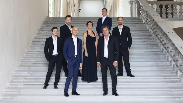 Eine Gruppe elegant gekleideter Menschen (Accademia Bizantina mit Ottavio Dantone) steht auf einer breiten Marmortreppe in einem historischen Gebäude. Sie tragen formelle Kleidung.